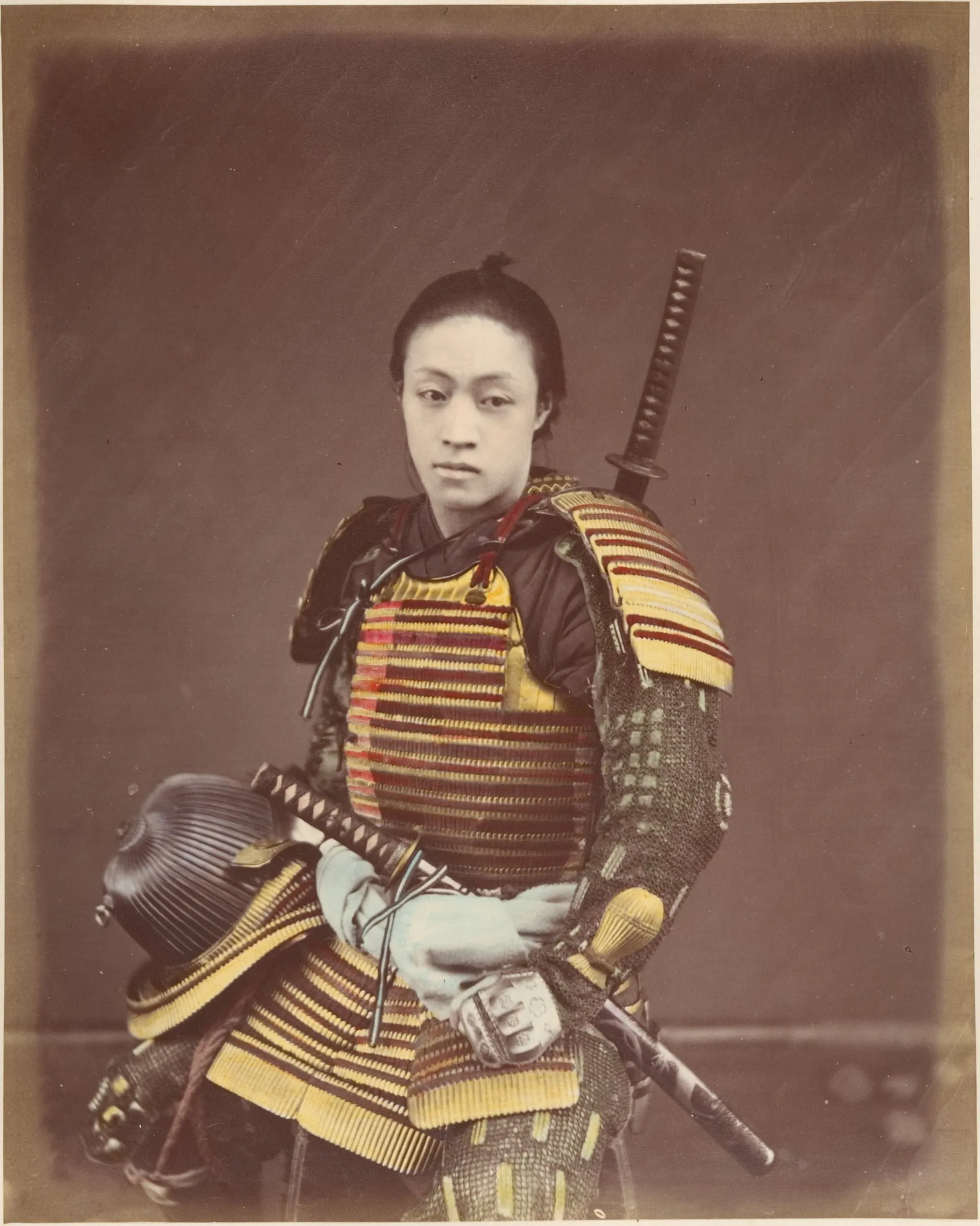 Fotografía coloreada a mano de un joven con coraza de láminas amarillas y rojas, con una espada atrás y otra en la mano.