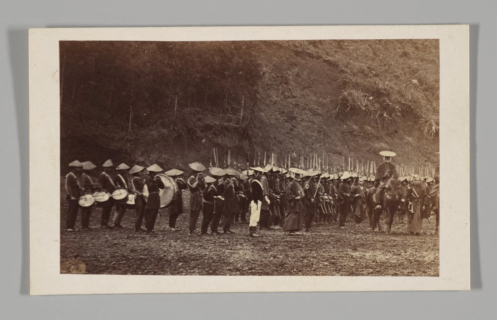 Fotografía antigua de una larga fila de hombres con sombreros cónicos, fusiles y tambores, en un camino de tierra.