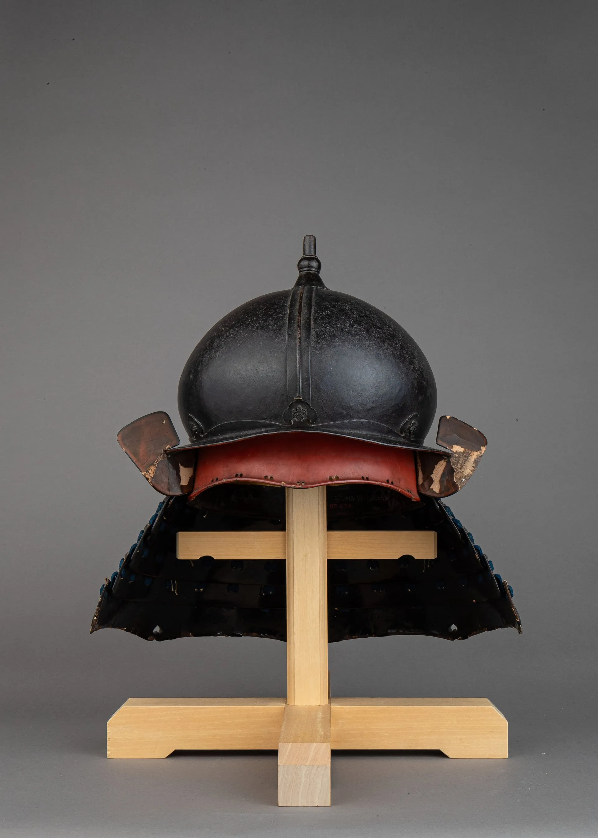 Casco de hierro negro con cubrenuca de láminas, sobre un soporte de madera clara.