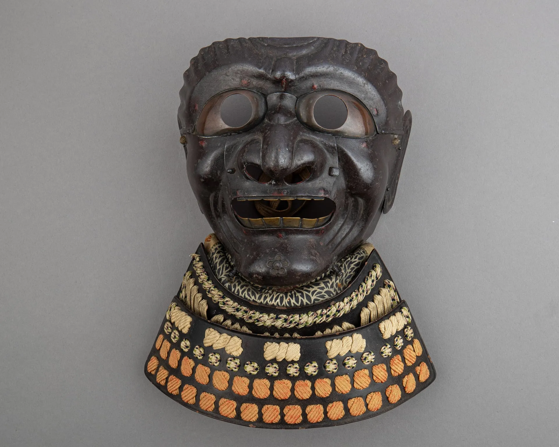Máscara de hierro oscuro con rasgos de rostro humano, dientes de metal y una gola de láminas con cordones.
