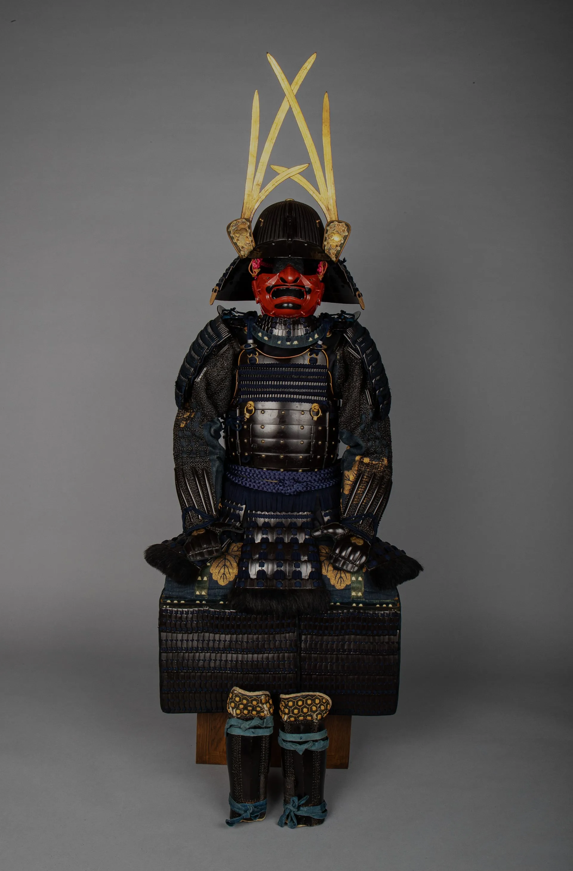 Armadura completa sobre un soporte: yelmo negro con una cimera dorada, máscara roja y coraza de láminas oscuras.