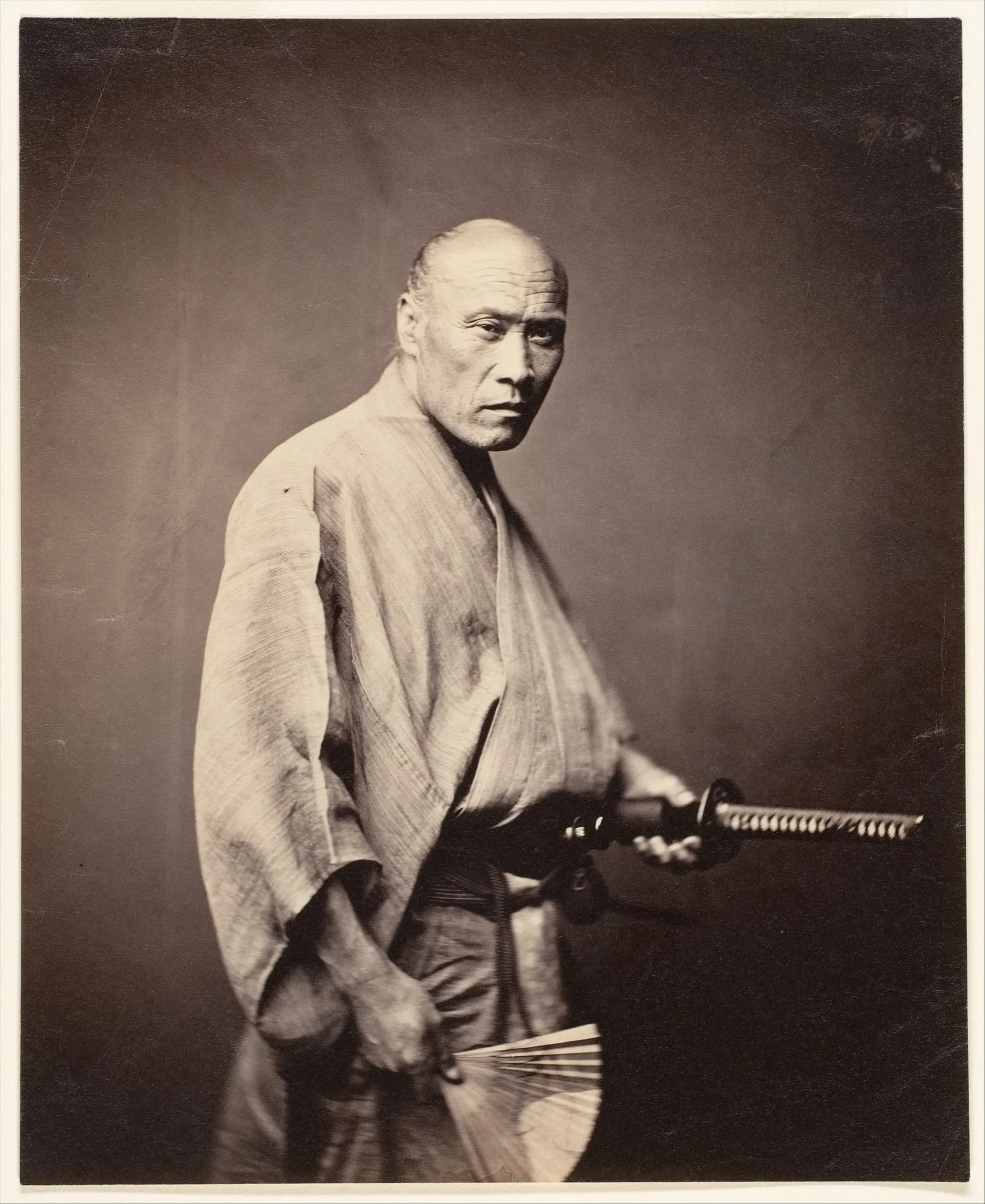 Fotografía antigua de un hombre de pie, con kimono claro, una espada al cinto y un abanico en la mano.