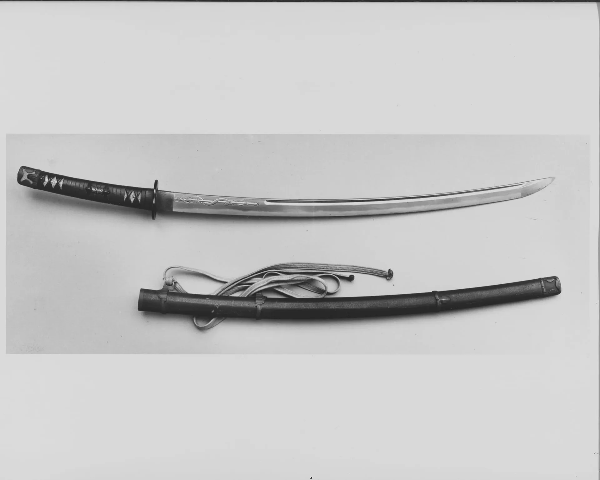 Fotografía en blanco y negro de una espada curva con su vaina y un cordón enrollado.